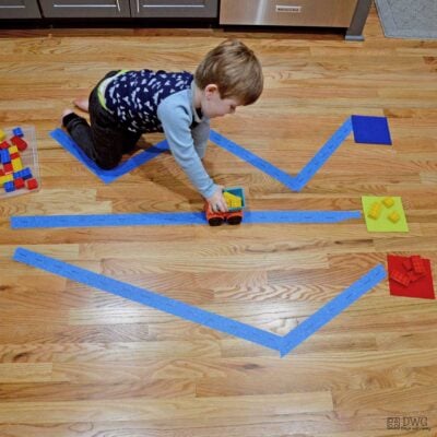 Preschooler sorting yellow Duplos in a toy truck to a yellow square. He is following a road made from painter's tape.