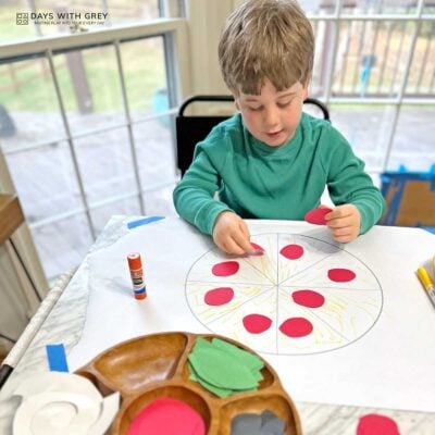Preschooler sitting at a table decorating a paper pizza with red paper circles for pepperoni.