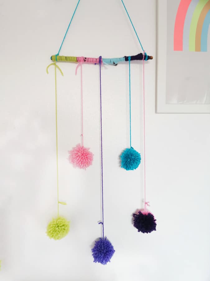 yarn wrapped stick and pom pom mobile against white wall