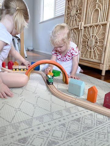 Two little girls playing with a wooden train set and a Grimms wooden rainbow.