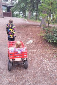 Older girl exploring nature while pulling a baby in a red wagon on a woodland path