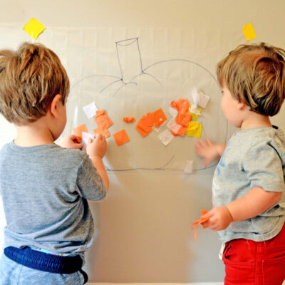Two toddlers adding yellow and orange tissue paper squares to contact paper hung on the wall. The sticky paper has a pumpkin outline that the children are decorating for fall.