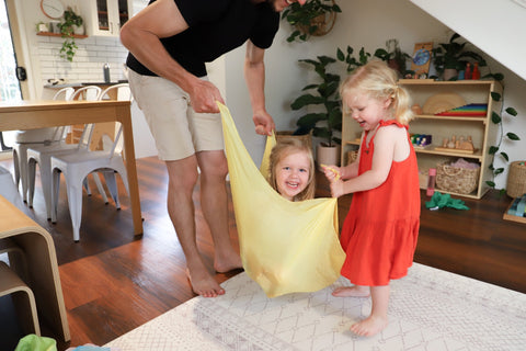 A family playing with a large yellow Sarah's Silk play silk. The youngest daughter is sitting in the silk and being lifted up.