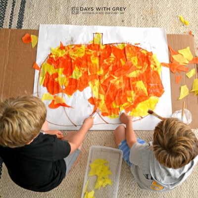 Two boys sitting on the kitchen floor working on a pumpkin art activity. They are gluing pieces of orange and yellow tissue paper to the large drawing.