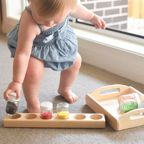 Toddler playing with small glass jars in a wooden tray with coloured rice.