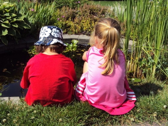 preschool boy red shirt, preschool girl pink shirt, sitting beside backyard pond