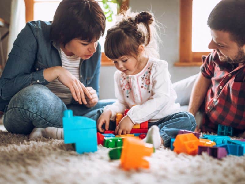 A young girl sits on the floor with her mom and dad during free play time, playing with some plastic building blocks while her parents watch.