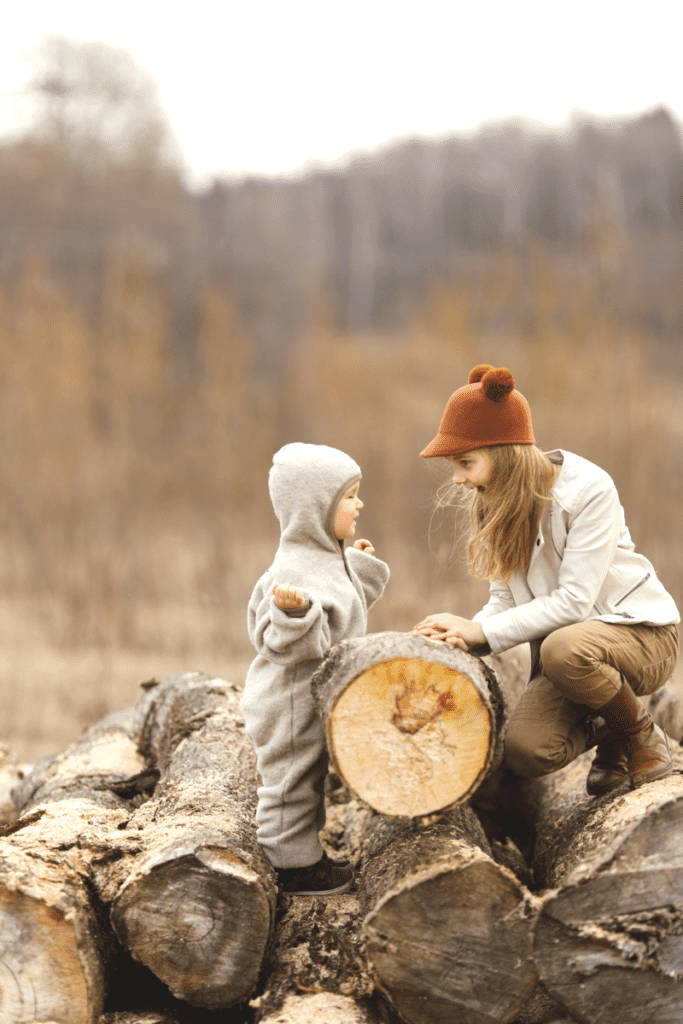 A toddler standing on a log while looking at a woman. Both are smiling while they do an outdoor activity.